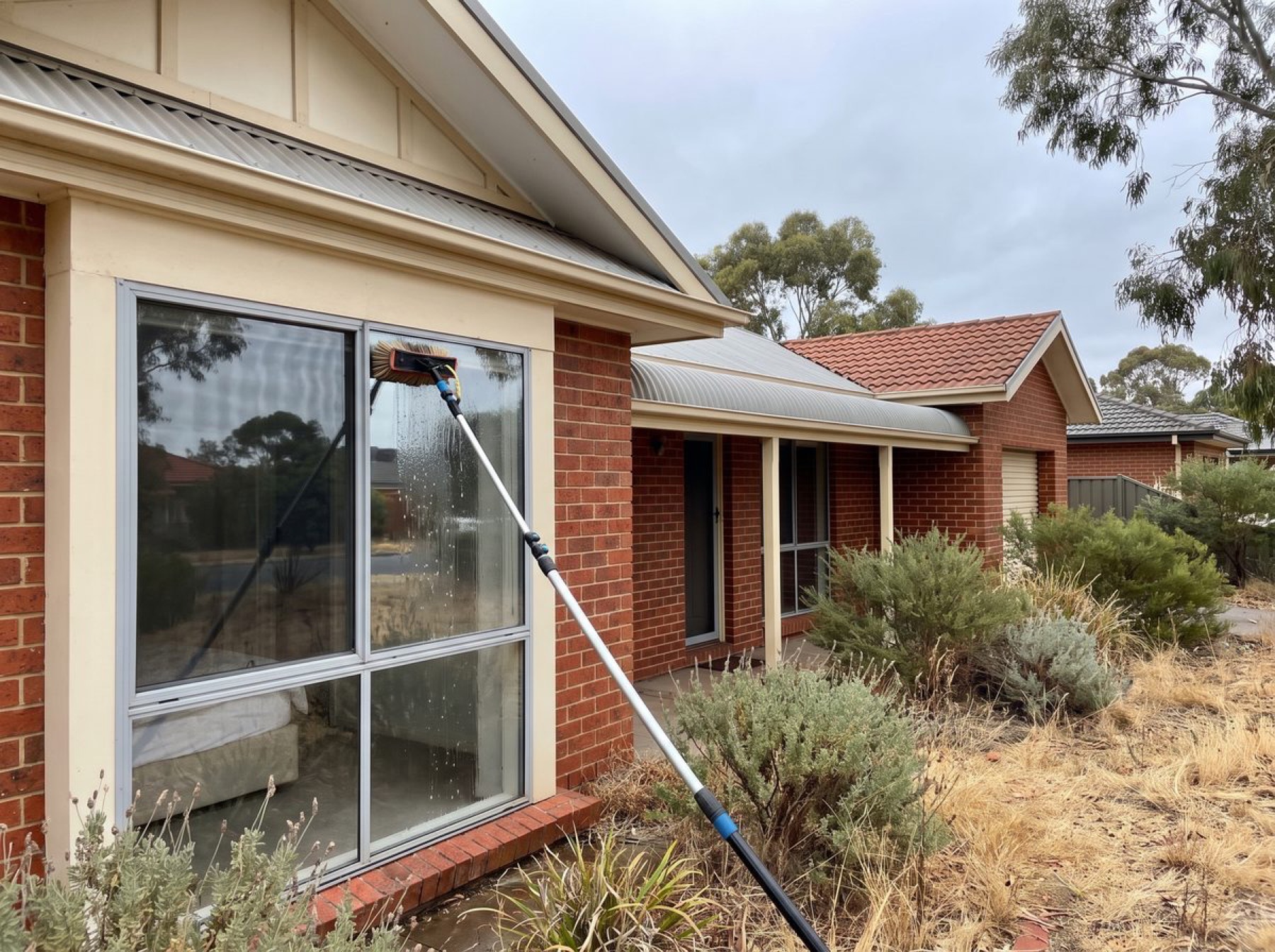 A person using a long-handled brush to clean a window at a brick house