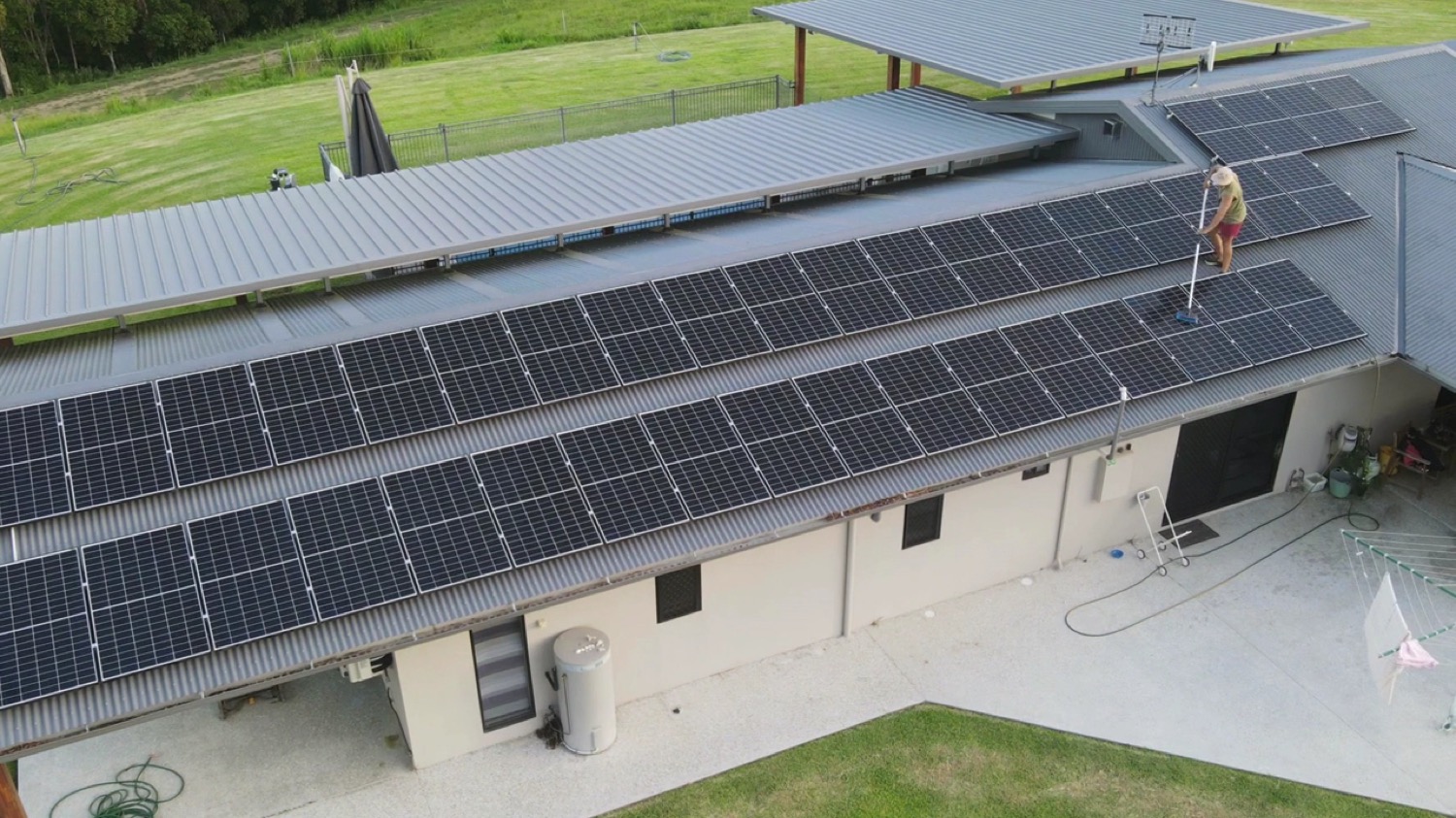 A worker using a long-handled brush to clean solar panels on a large metal roof